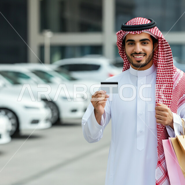 Expressions of joy and happiness with electronic payment operations, discounts and offers in Saudi Arabia, a smiling Saudi Arabian Gulf man wearing a traditional shemagh and thobe, holding colorful paper bags and a credit card, the joy of shopping and purchasing supplies and gifts from shops and malls