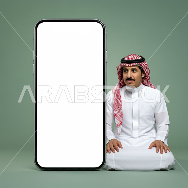 النظر إلى جهاز الجوال بايماءات الصدمة والذهول، بورتريه لرجل عربي خليجي سعودي يرتدي الثوب التقليدي والشماغ يجلس أمام مجسم ضخم لهاتف محمول بشاشة بيضاء فارغة، الإعلانات والعروض التسويقية عبر التقنيات والتكنولوجيا الحديثة، موك أب أبيض كبير، خلفية خضراء