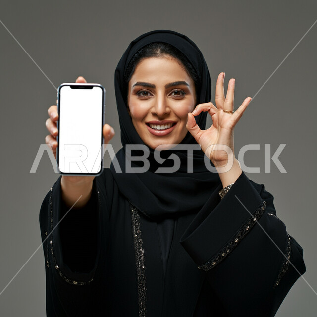 النظر الى الكاميرا بتعابير القبول والفرح والسرور، الإعلانات والعروض التسويقية، بورتريه مقرب لإمرأة عربية خليجية سعودية محجبة ترتدي العباءة ترفع يدها للأعلى بإيماءات الإعجاب والرضا، علامة الجودة والامتياز، عرض شاشة بيضاء فارغة على الجوال، خلفية رمادية