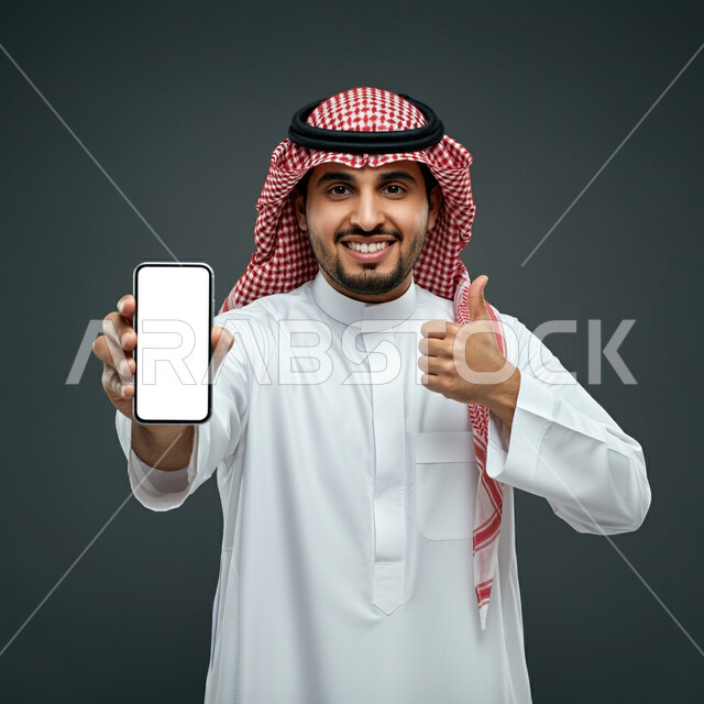 Looking at the camera with expressions of acceptance, joy and pleasure, advertisements and marketing offers, close-up portrait of a Saudi Arabian Gulf man wearing traditional thobe and shemagh raising index finger with gestures of admiration and satisfaction, quality and excellence mark, displaying a blank white screen on a mobile phone, gray background
