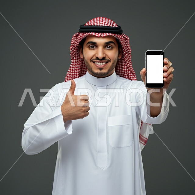النظر الى الكاميرا بتعابير القبول والفرح والسرور، الإعلانات والعروض التسويقية، بورتريه مقرب لرجل عربي خليجي سعودي يرتدي الثوب التقليدي والشماغ يرفع اصبع السبابة بإيماءات الإعجاب والرضا، علامة الجودة والامتياز، عرض شاشة بيضاء فارغة على الجوال، خلفية رمادية