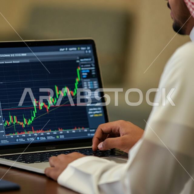 استخدام الأجهزة التقنية الحديثة، صورة مقربة من الجانب لرجل أعمال عربي خليجي سعودي يرتدي الثوب التقليدي والشماغ يقوم بالعمل على الحاسوب المحمول، مراقبة الرسوم البيانية الهولوجرامية على الشاشات الإلكترونية المضيئة، متابعة ارتفاع وانخفاض الأسهم في سوق البورصة