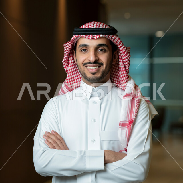 الوقوف بايماءات أيدي متقاطعة، الاناقة والاهتمام بالمظهر الخارجي، صورة مقربة لرجل عربي خليجي سعودي مبتسم يرتدي الثوب التقليدي والشماغ ينظر الى الكاميرا بتعابير الفخر والثقة بالنفس، مفهوم الشياكة والرجولة والقوة