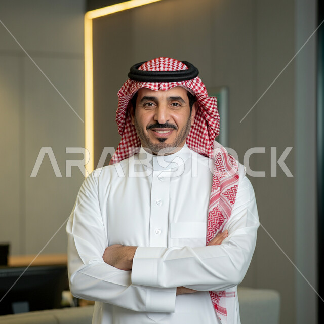 الوقوف بايماءات أيدي متقاطعة، الاناقة والاهتمام بالمظهر الخارجي، صورة مقربة لرجل عربي خليجي سعودي مبتسم يرتدي الثوب التقليدي والشماغ ينظر الى الكاميرا بتعابير الفخر والثقة بالنفس، مفهوم الشياكة والرجولة والقوة