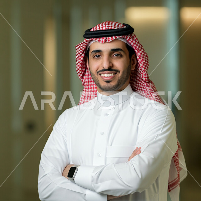الوقوف بايماءات أيدي متقاطعة، الاناقة والاهتمام بالمظهر الخارجي، صورة مقربة لرجل عربي خليجي سعودي مبتسم يرتدي الثوب التقليدي والشماغ ينظر الى الكاميرا بتعابير الفخر والثقة بالنفس، مفهوم الشياكة والرجولة والقوة