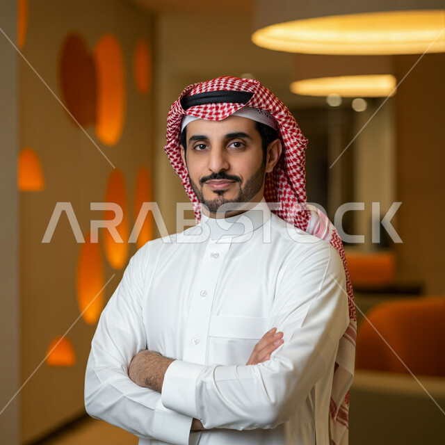 الوقوف بايماءات أيدي متقاطعة، الاناقة والاهتمام بالمظهر الخارجي، صورة مقربة لرجل عربي خليجي سعودي مبتسم يرتدي الثوب التقليدي والشماغ ينظر الى الكاميرا بتعابير الفخر والثقة بالنفس، مفهوم الشياكة والرجولة والقوة