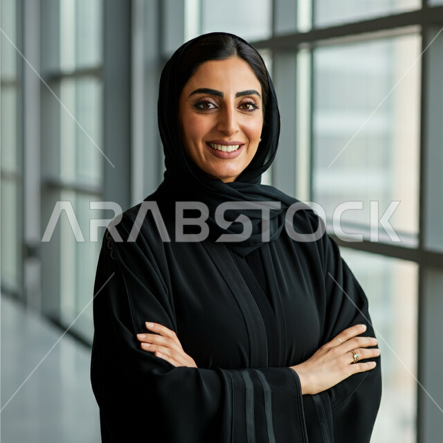 The concept of femininity and softness, interest in elegance and external appearance, a close-up image of a smiling Saudi Arabian Gulf woman wearing a black abaya looking at the camera with expressions of pleasure and self-confidence, standing with crossed hand gestures