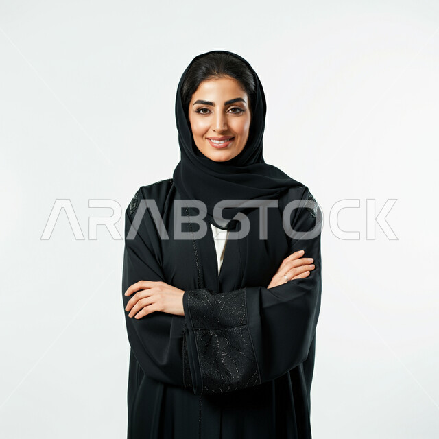 The concept of femininity and softness, interest in elegance and external appearance, portrait of a smiling Saudi Arabian Gulf woman wearing a black abaya looking at the camera with expressions of pleasure and self-confidence, standing with crossed hand gestures, white background