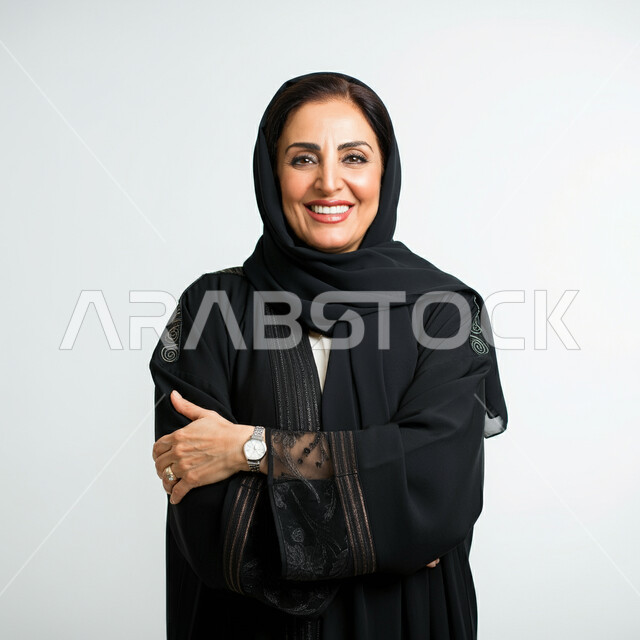 مفهوم الانوثة والنعومة، الاهتمام بالاناقة والمظهر الخارجي،  بورتريه لامرأة عربية خليجية سعودية مبتسمة ترتدي العباءة السوداء تنظر الى الكاميرا بتعابير السرور والثقة بالنفس، الوقوف بايماءات ايدي متقاطعة، خلفية بيضاء