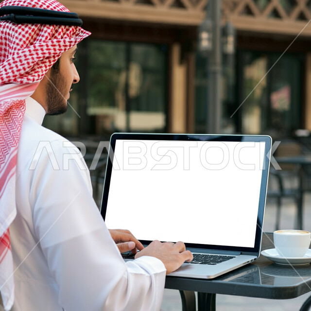 تصفح التطبيقات والبرامج الالكترونية عبر الابتوب, استخدام الأجهزة التقنية الحديثة والمتطورة, إدراة الأعمال عن بعد, رجل عربي خليجي سعودي يرتدي الثوب التقليدي والشماغ يعمل على جهاز الحاسب المحمول، عرض شاشة بيضاء فارغة، الاستمتاع بالمشروبات الساخنة في أحد المقاهي