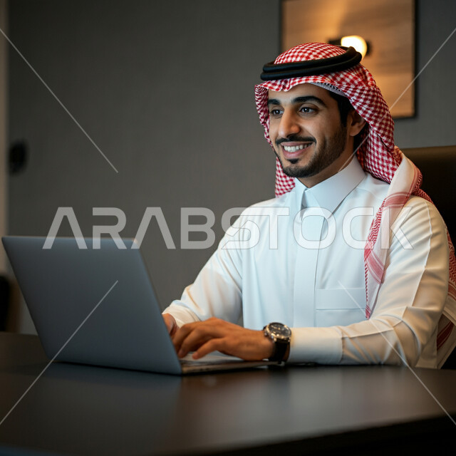 أداء المهام والاعمال المكتبية داخل مقر الشركة، رجل عربي خليجي سعودي مبتسم يرتدي الثوب التقليدي والشماغ يجلس أمام مكتبه يقوم بالعمل على الحاسب المحمول، دمج التقنية والتكنولوجيا في الحياة العملية، العمل في مكاتب وشركات المملكة العربية السعودية