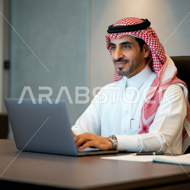 Performing office tasks and work inside the company headquarters, a smiling Saudi Arabian Gulf man wearing the traditional thobe and shemagh sitting in front of his desk working on the laptop, integrating technology and technology into practical life, working in offices and companies in the Kingdom of Saudi Arabia
