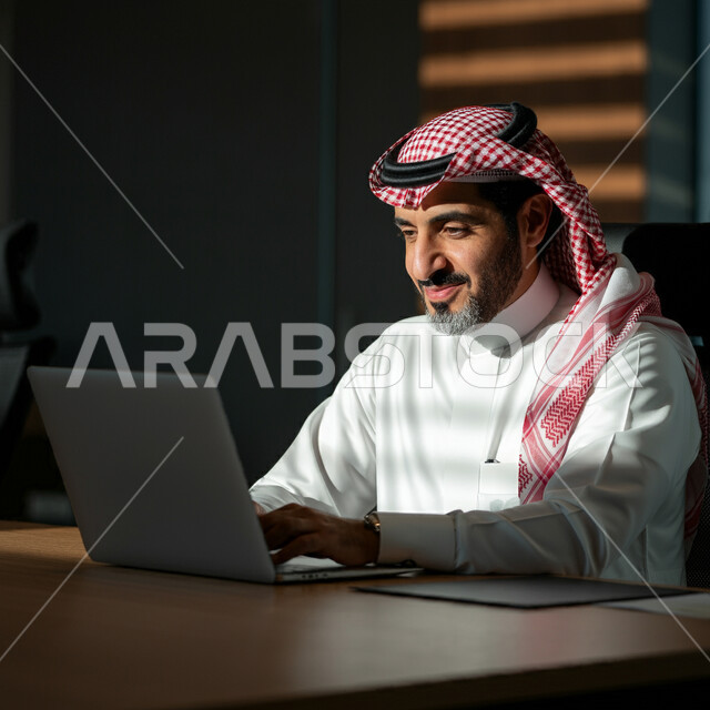 أداء المهام والاعمال المكتبية داخل مقر الشركة، رجل عربي خليجي سعودي مبتسم يرتدي الثوب التقليدي والشماغ يجلس أمام مكتبه يقوم بالعمل على الحاسب المحمول، دمج التقنية والتكنولوجيا في الحياة العملية، العمل في مكاتب وشركات المملكة العربية السعودية