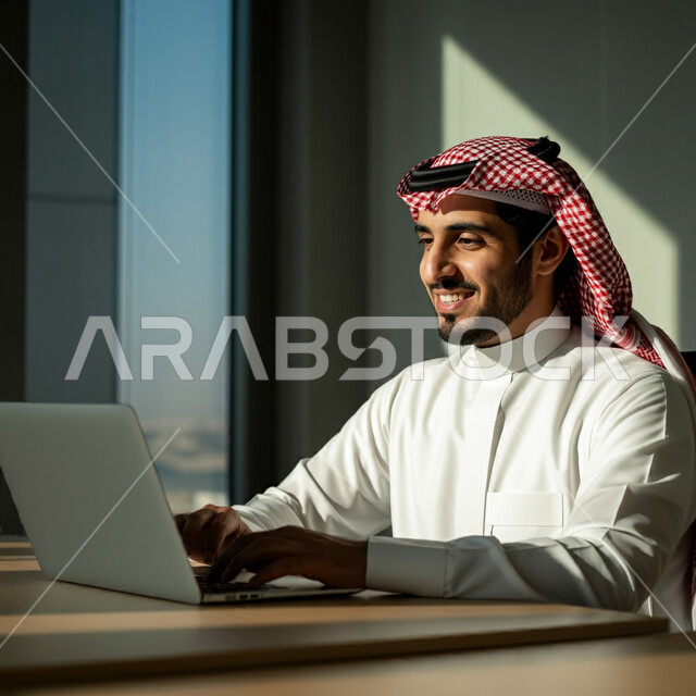 أداء المهام والاعمال المكتبية داخل مقر الشركة، رجل عربي خليجي سعودي مبتسم يرتدي الثوب التقليدي والشماغ يجلس أمام مكتبه يقوم بالعمل على الحاسب المحمول، دمج التقنية والتكنولوجيا في الحياة العملية، العمل في مكاتب وشركات المملكة العربية السعودية