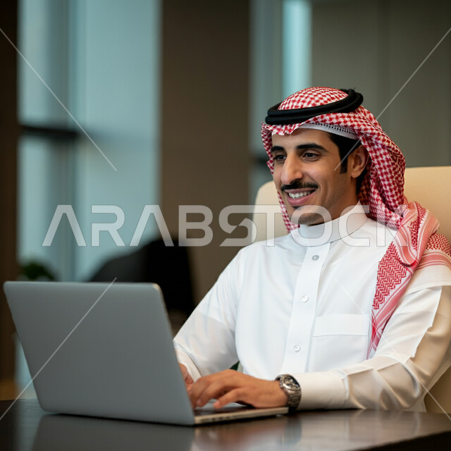 أداء المهام والاعمال المكتبية داخل مقر الشركة، رجل عربي خليجي سعودي مبتسم يرتدي الثوب التقليدي والشماغ يجلس أمام مكتبه يقوم بالعمل على الحاسب المحمول، دمج التقنية والتكنولوجيا في الحياة العملية، العمل في مكاتب وشركات المملكة العربية السعودية