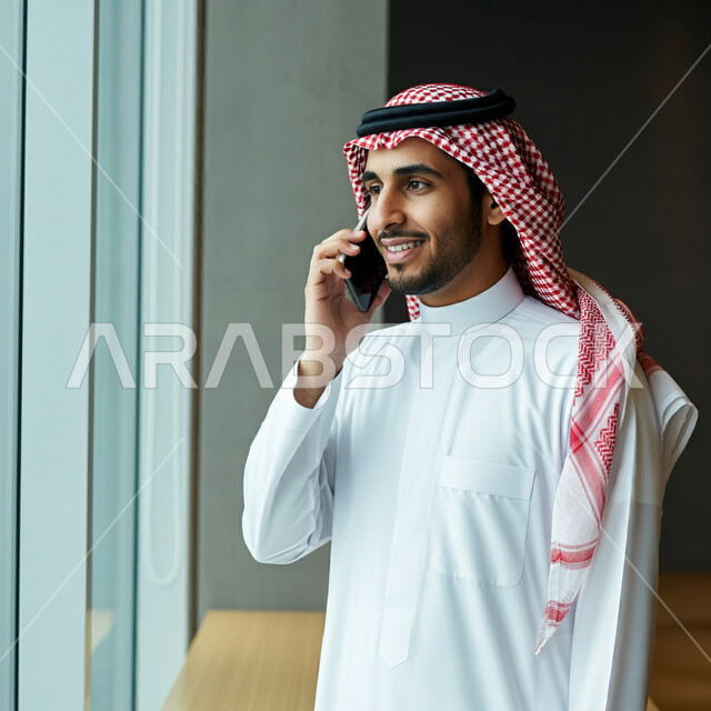 دمج التقنية والتكنولوجيا في الحياة اليومية والعملية، رجل عربي خليجي سعودي مبتسم يرتدي الثوب التقليدي والشماغ يقوم بأجراء مكالمة هاتفية باستخدام الهاتف المحمول داخل مقر الشركة، التواصل مع الاهل والاصدقاء، ايماءات الفرح والسرور
