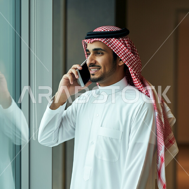 دمج التقنية والتكنولوجيا في الحياة اليومية والعملية، رجل عربي خليجي سعودي مبتسم يرتدي الثوب التقليدي والشماغ يقوم بأجراء مكالمة هاتفية باستخدام الهاتف المحمول داخل مقر الشركة، التواصل مع الاهل والاصدقاء، ايماءات الفرح والسرور