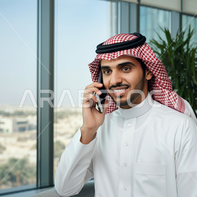 دمج التقنية والتكنولوجيا في الحياة اليومية والعملية، رجل عربي خليجي سعودي مبتسم يرتدي الثوب التقليدي والشماغ يقوم بأجراء مكالمة هاتفية باستخدام الهاتف المحمول داخل مقر الشركة، التواصل مع الاهل والاصدقاء، ايماءات الفرح والسرور