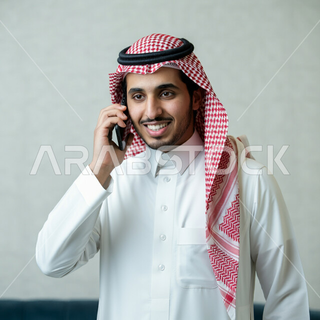 Gestures of joy and happiness to hear good news, integrating technology and technology into daily and practical life, close-up portrait of a smiling Saudi Arabian Gulf man wearing traditional thobe and shemagh making a phone call using a mobile phone, communicating with family and friends, white background