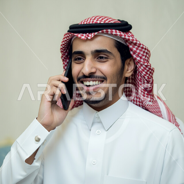  ايماءات الفرح والسرور لسماع الاخبار المفرحة، دمج التقنية والتكنولوجيا في الحياة اليومية والعملية، بورتريه مقرب لرجل عربي خليجي سعودي مبتسم يرتدي الثوب التقليدي والشماغ يقوم بأجراء مكالمة هاتفية باستخدام الهاتف المحمول، التواصل مع الاهل والاصدقاء، خلفية بيضاء