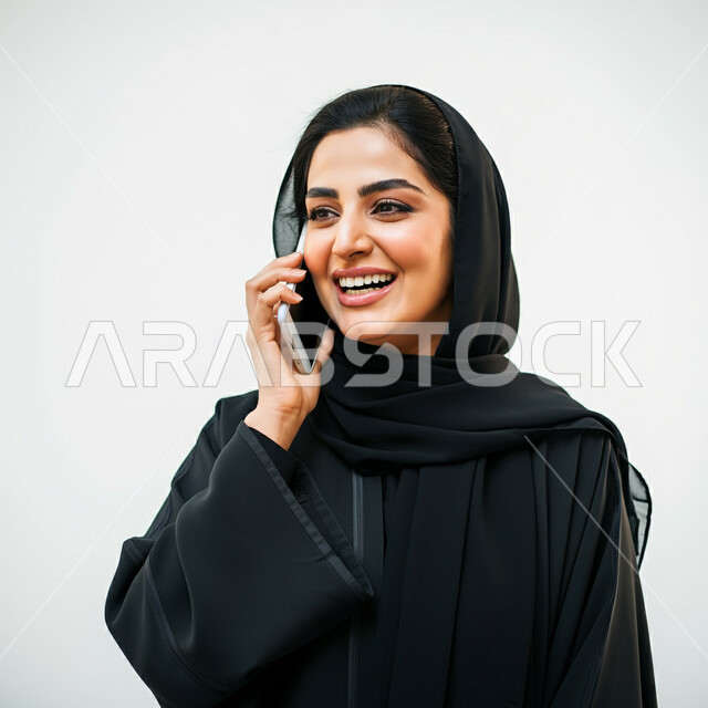التواصل مع الاهل والاصدقاء، ايماءات الفرح والسرور لسماع الاخبار المفرحة، دمج التقنية والتكنولوجيا في الحياة اليومية، بورتريه مقرب لامرأة عربية خليجية سعودية مبتسمة ترتدي العباءة السوداء تقوم بأجراء مكالمة هاتفية باستخدام الجوال، دردشة ومحادثات وتعارف، خلفية بيضاء
