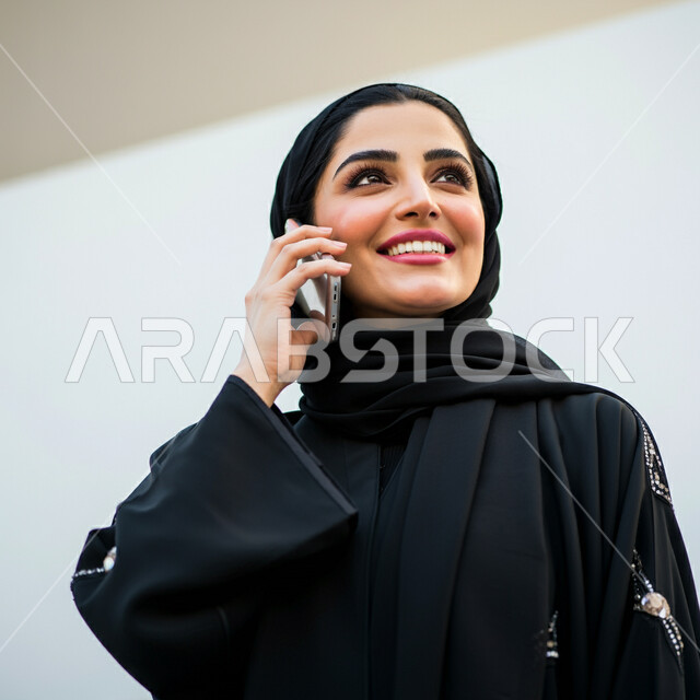 التواصل مع الاهل والاصدقاء، ايماءات الفرح والسرور لسماع الاخبار المفرحة، دمج التقنية والتكنولوجيا في الحياة اليومية، بورتريه مقرب لامرأة عربية خليجية سعودية مبتسمة ترتدي العباءة السوداء تقوم بأجراء مكالمة هاتفية باستخدام الجوال، دردشة ومحادثات وتعارف، خلفية بيضاء