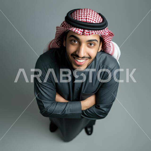 الاهتمام بالأناقة و المظهر الخارجي، مفهوم الرجولة والثقة بالنفس، بورتريه من الاعلى لرجل عربي خليجي سعودي يرتدي الشماغ والثوب التقليدي يقف بأيدي متقاطعة ينظر الى الكاميرا بتعابير السرور, الوقوف باستعداد واستقامة، خلفية رمادية