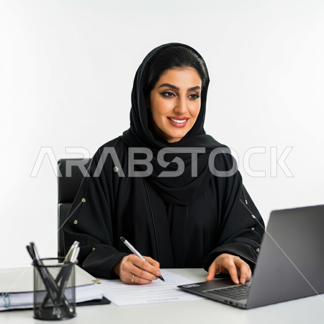 أستخدام التقنيات والحاسب في تنظيم وادارة الاعمال، بورتريه مقرب لامرأة عربية خليجية سعودية مبتسمة ترتدي العباءة تجلس امام مكتبها تقوم بتسجيل وتدوين الملاحظات والمعلومات على الورق، العمل في مكاتب وشركات المملكة العربية السعودية، مهن و وظائف نسائية، خلفية بيضاء