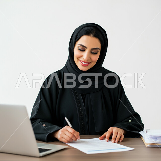 أستخدام التقنيات والحاسب في تنظيم وادارة الاعمال، بورتريه مقرب لامرأة عربية خليجية سعودية مبتسمة ترتدي العباءة تجلس امام مكتبها تقوم بتسجيل وتدوين الملاحظات والمعلومات على الورق، العمل في مكاتب وشركات المملكة العربية السعودية، مهن و وظائف نسائية، خلفية بيضاء