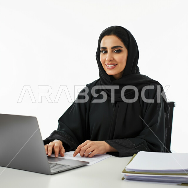 العمل في مكاتب وشركات المملكة العربية السعودية، أستخدام التقنيات والتكنولوجيا في تنظيم وادارة الاعمال، بورتريه لامرأة عربية خليجية سعودية مبتسمة ترتدي العباءة تجلس امام مكتبها تقوم باستخدام جهاز اللابتوب، مهن و وظائف نسائية مكتبية، خلفية بيضاء