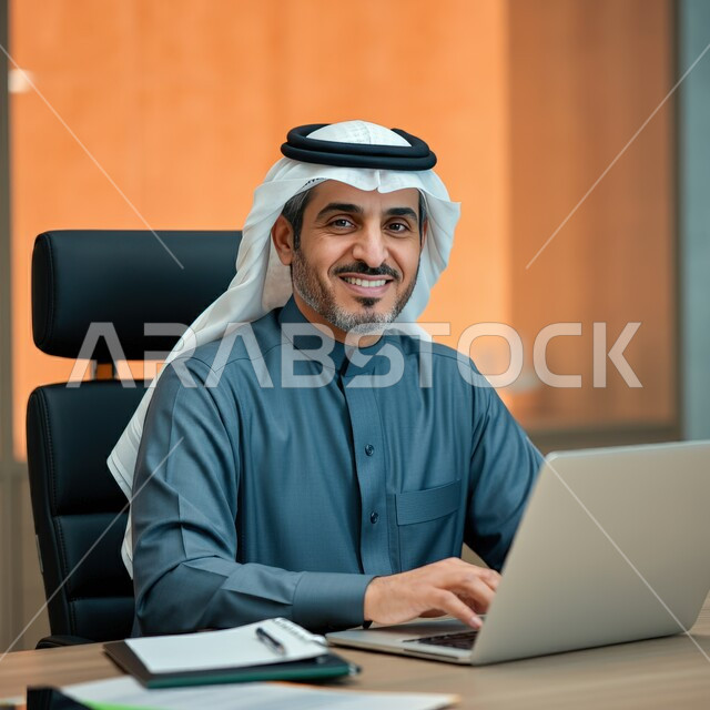 أداء المهام والاعمال المكتبية داخل مقر الشركة، رجل عربي خليجي سعودي مبتسم يرتدي الثوب التقليدي والشماغ يجلس أمام مكتبه يقوم بالعمل على الحاسب المحمول ينظر الى الكاميرا بإيماءات السرور، دمج التقنية والتكنولوجيا في الحياة العملية، العمل في مكاتب المملكة