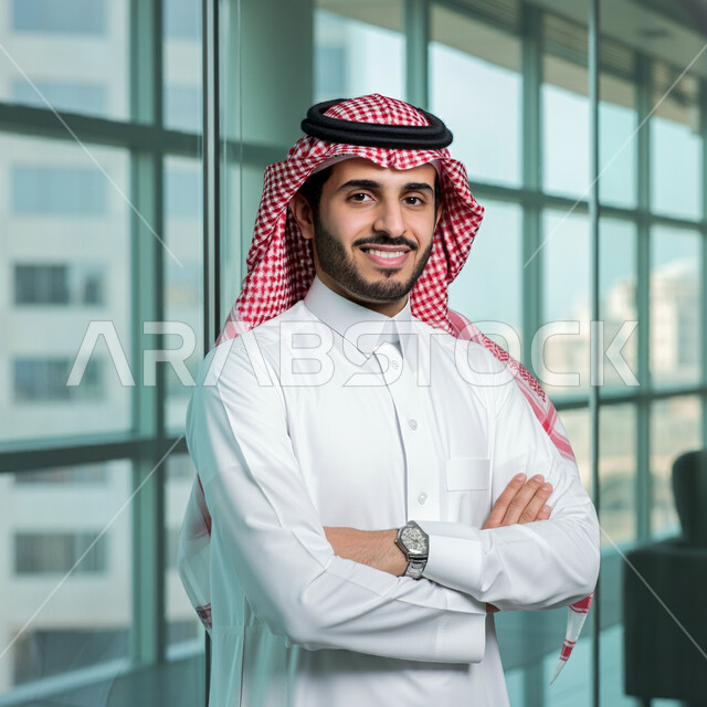 التقاط صور للوثايق الشخصية، الاناقة والاهتمام بالمظهر الخارجي، رجل عربي خليجي سعودي مبتسم يرتدي الثوب التقليدي والشماغ يقف بايماءات ايدي متقاطعة، النظر الى الكاميرا بتعابير الفرح والسرور، مفهوم القوة والشياكة والرجولة