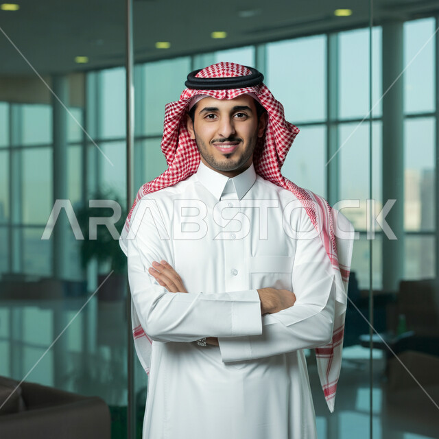 التقاط صور للوثايق الشخصية، الاناقة والاهتمام بالمظهر الخارجي، رجل عربي خليجي سعودي مبتسم يرتدي الثوب التقليدي والشماغ يقف بايماءات ايدي متقاطعة، النظر الى الكاميرا بتعابير الفرح والسرور، مفهوم القوة والشياكة والرجولة