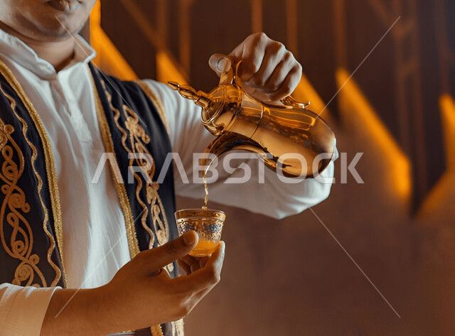 اتباع العادات والتقاليد السعودية في تقديم القهوة العربية بالدله النحاسية والفنجان، حسن الضيافه والإستقبال في الأعياد والمناسبات، منتجات وطنية محلية، تقديم المشروبات الساخنة، صورة مقربة لرجل عربي خليجي سعودي يرتدي الثوب التقليدي والدقلة يقوم بصب القهوة