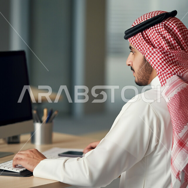 أداء المهام والاعمال المكتبية داخل مقر الشركة، صورة من الجانب لرجل عربي خليجي سعودي يرتدي الثوب التقليدي والشماغ  يجلس امام مكتبه يقوم بالعمل على الحاسب المحمول، دمج التقنية الحديثة والتكنولوجيا في الحياة العملية، العمل في مكاتب وشركات المملكة العربية السعودية