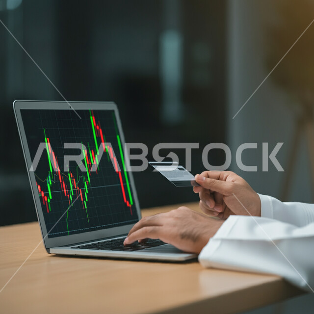 Economic investment concept, monitoring economy and financial information, close-up of a Saudi Arabian Gulf businessman wearing traditional thobe holding a credit card in his hand, following the rise and fall of stocks in the stock market through a laptop, using modern technical devices