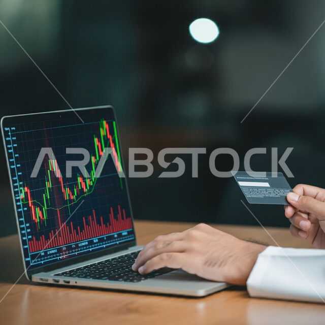استخدام الأجهزة التقنية الحديثة، مراقبة الاقتصاد والمعلومات المالية، صورة مقربة لرجل أعمال عربي خليجي سعودي يرتدي الثوب التقليدي يمسك بيده بطاقة الائتمان يقوم بمتابعة ارتفاع وانخفاض الأسهم في البورصة من خلال الحاسوب المحمول، مفهوم الاستثمار الاقتصادي