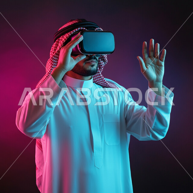 الاندماج ومحاكاة العالم الخيالي، بورتريه لرجل عربي خليجي سعودي يرتدي الثوب التقليدي والشماغ يضع نظارة الواقع الافتراضي يرفع يديه للاعلى بايماءات صد شيء ما والدفاع عن النفس، دمج التسلية بالتقنية والتكنولوجيا، قضاء اوقات ممتعة في عطلة نهاية الاسبوع، خلفية ملونة