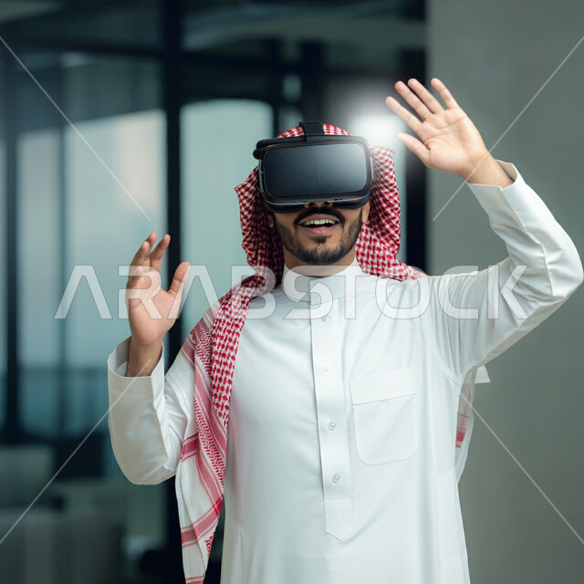 الاندماج ومحاكاة العالم الخيالي، لرجل عربي خليجي سعودي يرتدي الثوب التقليدي والشماغ يضع نظارة الواقع الافتراضي يرفع يديه للاعلى بايماءات صد شيء ما والدفاع عن النفس، دمج التسلية بالتقنية والاجهزة الحديثة والتكنولوجيا، قضاء اوقات ممتعة في عطلة نهاية الاسبوع