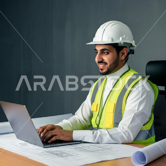 دمج التكنولوجيا والأجهزة الحديثة بالأعمال، مخططات البناء على طاولة مكتبية، بورتريه مقرب لمهندس عربي خليجي سعودي يرتدي سترة الحماية والخوذة يقوم باستخدام الحاسوب المحمول, إنجاز مهام العمل ومتابعة سير المشروع، وظائف شبابية في المملكة العربية السعودية، خلفية رمادية