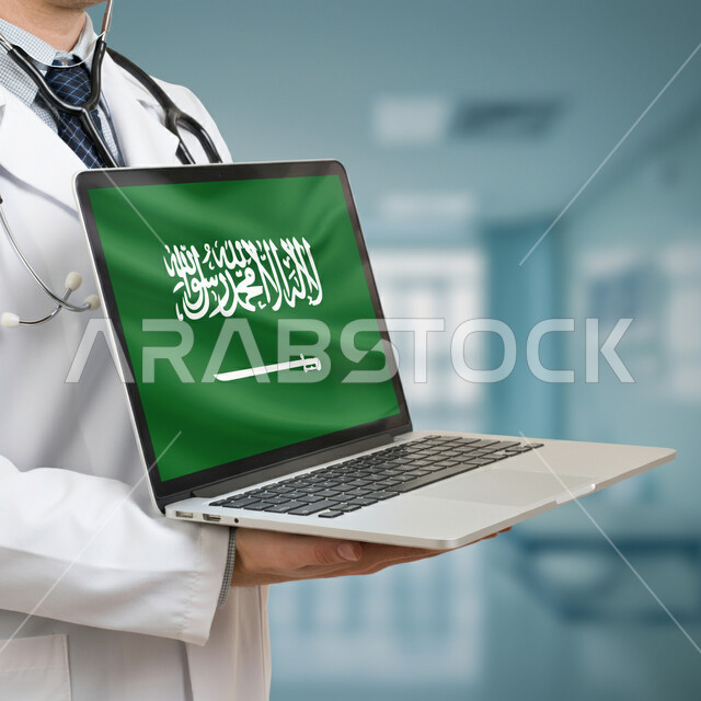 استخدام الأجهزة الالكترونية الحديثة في المجال الطبي، عرض علم المملكة العربية السعودية على شاشة الحاسوب المحمول في اليوم الوطني السعودي 23 سبتمبر، صورة مقربة لطبيب عربي خليجي سعودي يرتدي المعطف الطبي يقوم بتقديم الرعاية الصحية لأبناء الوطن
