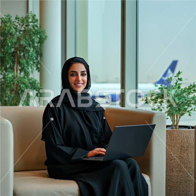 انجاز مهام الاعمال عن بعد، مفهوم الرحلات الجوية السياحية, سيدة أعمال عربية سعودية خليجية محجبة ترتدي العباءة السوداء تجلس داخل صالة الانتظار في المطار تعمل على جهاز الحاسب المحمول, الاستعداد للسفر في رحلة عمل ممتعة, النظر الى الكاميرا بايماءات السعادة والحماس