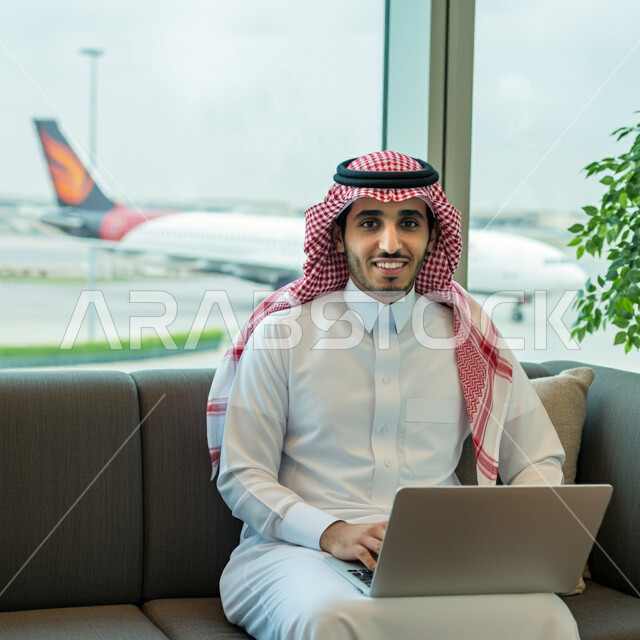 إنجاز العمل من خلال جهاز اللابتوب, قضاء عطلة صيفية ممتعة، مفهوم الرحلات الجوية السياحية, رجل أعمال عربي سعودي خليجي مبتسم يرتدي الثوب التقليدي داخل صالة الانتظار في المطار, الاستعداد للسفر في رحلة عمل, أهمية السياحة في المملكة العربية السعودية