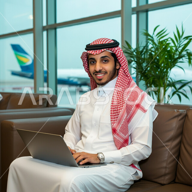 قضاء عطلة صيفية ممتعة، مفهوم الرحلات الجوية السياحية, رجل أعمال عربي سعودي خليجي مبتسم يرتدي الثوب التقليدي التقليدي داخل صالة الانتظار في المطار, إنجاز العمل من خلال جهاز اللابتوب, الاستعداد للسفر في رحلة عمل, أهمية السياحة في المملكة العربية السعودية