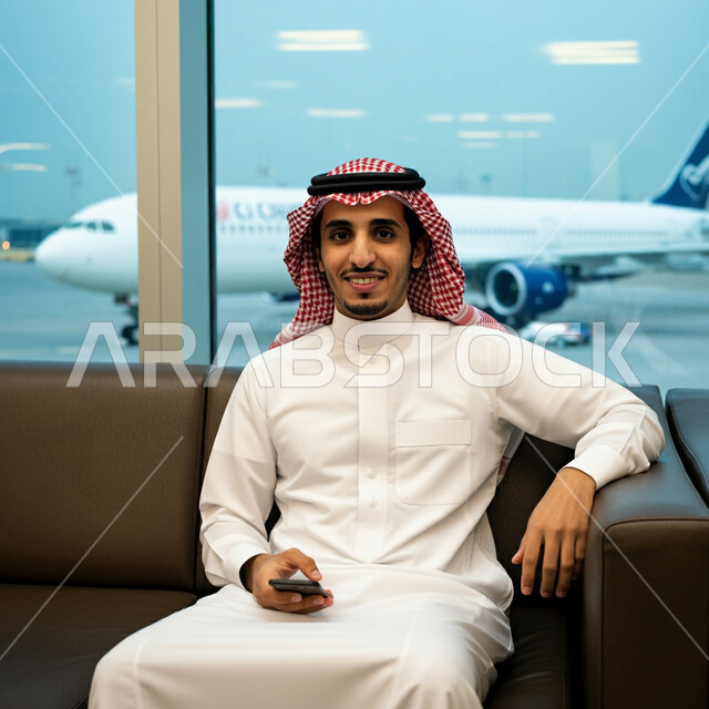 انتظار موعد إقلاع الطائرة, رجل أعمال عربي سعودي خليجي مبتسم يرتدي الثوب التقليدي والشماغ يجلس داخل قاعة الانتظار في المطار ويحمل بيده الهاتف المحمول، النظر الى الكاميرا بايماءات السعادة، مفهوم السياحة والسفر