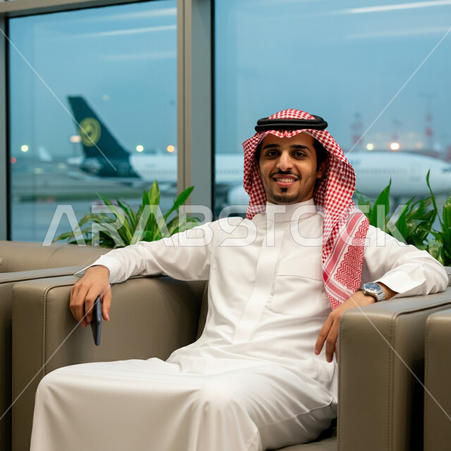 Sharing trip details on social media, browsing the internet and following the news, a smiling Saudi Arabian Gulf businessman wearing a Saudi thobe and a shemagh sitting inside the airport waiting room holding a mobile phone, looking at the camera with gestures of happiness, the concept of tourism and travel