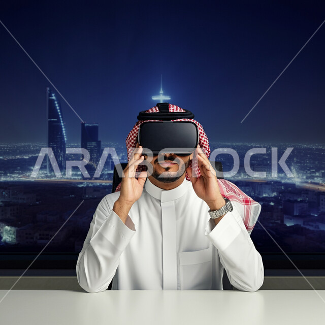 الإندماج ومحاكاة العالم الخيالي، المتعة والتسلية بالمشاهدة عبر نظارة الواقع الافتراضي، رجل عربي خليجي سعودي يرتدي الثوب التقليدي والشماغ يجلس أمام مكتبه يضع نظارات ثلاثية الأبعاد، استخدام التقنية والاجهزة الحديثة، خلفية الابراج وناطحات السحاب
