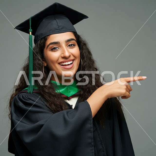 تحديد موقع شيء ما، الاحتفال بإنهاء الدراسة الجامعية، الشعور بالفخر والثقة بالنفس، مفهوم الاجتهاد والتفوق، بورتريه مقرب لطالبة عربية خليجية سعودية ترتدي عباءه وقبعة التخرج تشير لمكان ما بايماءات السرور، خلفية رمادية