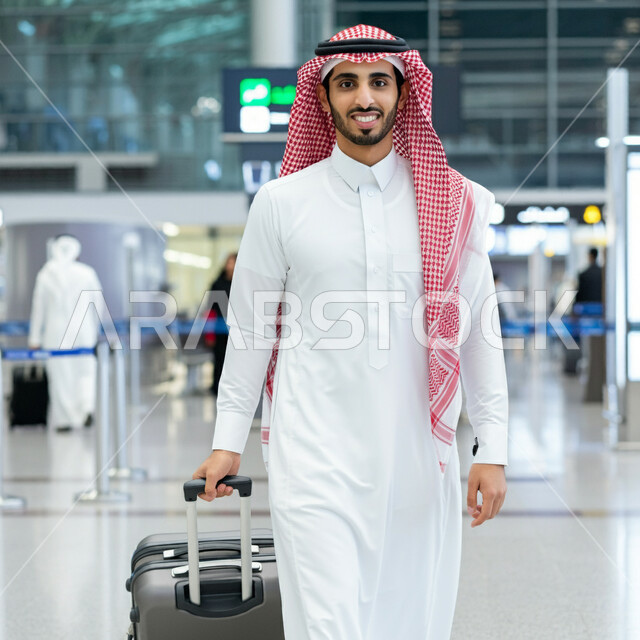 الاستعداد للذهاب في رحلة ترفيهية، رجل عربي خليجي سعودي يرتدي الشماغ والثوب التقليدي يمسك بيده حقيبة الامتعة ينظر إلى الكاميرا بإيماءات السعادة والسرور، صالة الانتظار في مطارات المملكة العربية السعودية، قضاء إجازة صيفية ممتعة، الاناقة والاهتمام بالمظهر الخارجي