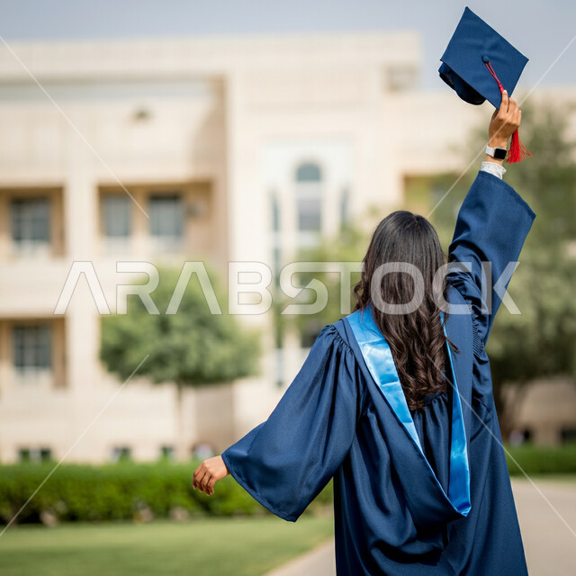 الاحتفال بإنهاء الدراسة الجامعية، مفهوم الاجتهاد والتفوق، الشعور بالفخر والثقة بالنفس، صورة مقربة من الخلف لطالبة عربية خليجية سعودية ترتدي عباءه التخرج تحمل بيدها القبعة